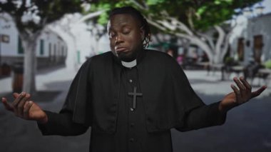 Papaz tasması ve çapraz kolyesi olan bir rahip, sokakta omuz silkme hareketinde avuç içlerini kaldırır..