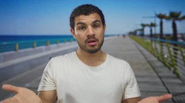 Açık elleri olan genç İspanyol adam, güneş gören sahil kıyısında palmiye ağaçlarıyla kaplı bir kaldırımda sürpriz yapar..