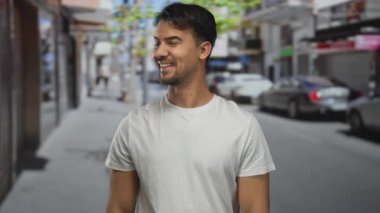 Sokak ortasında gülümseyen İspanyol genç bir adam güneşli bir günde şehir çevresiyle çevrili, güneşli bir arka planda arabaları ve dükkanları olan günlük beyaz gömlekler giyiyor.