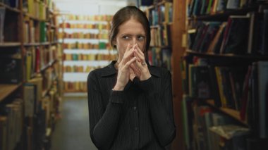 Yüzü kütüphane koridorunda endamlı ellere sahip bir kadın, uzun kitaplıklar arasında, sıcak ışık altında yan yan bakıyor..