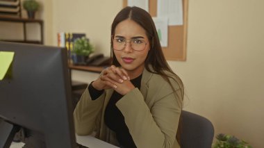 Modern ofiste masasında gözlüklü bir kadın dikkatle bilgisayara odaklanıyor, bitkilerle çevrili ve düzenli çalışma alanı yaratıyor profesyonel bir atmosfer yaratıyor..