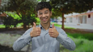 Genç İspanyol adam, yapraklı ağaçların altındaki güneşli yeşil parkta gülümserken iki kez baş parmak işareti yapıyor..