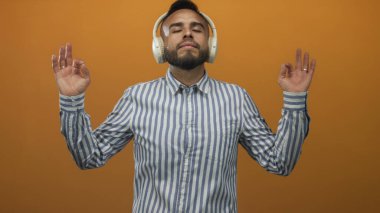 Kulaklık takan ve elleri çizgili gömlek giyen bir adam meditasyon yapıyor ve turuncu stüdyoda gözleri kapalı..