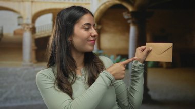 İspanyol kadın gülümsüyor ve arka planda kemerler olan eski bir kasabada bir zarfa işaret ediyor..