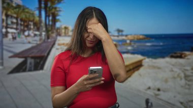 Woman pinching nose while holding smartphone on a street promenade by the seaside in a red shirt; discomfort.