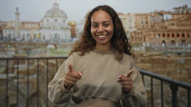 Parmaklarıyla gülümseyen bir kadın antik Roma binasında yıkıntılara ve parmaklıklara bakan kamerayı işaret ediyor; keyif yolculuğu keşfi.