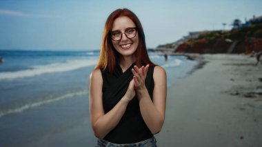 Gülümseyen kızıl saçlı kadın sahilde alkışlıyor, güneşli bir kumsal gününün keyfini yakalıyor dalgalar ve arka planda uzak uçurumlar.