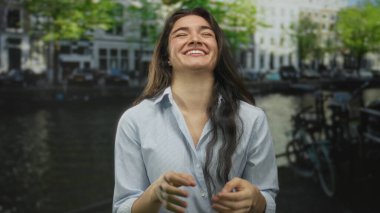 Young hispanic woman smiling and laughing with clasped hands wearing striped shirt on sunlit street; joy.