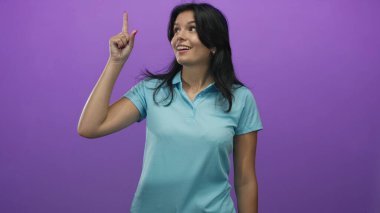 Mavi polo tişörtlü kadın mor stüdyoda parmak gösterip gülümsüyor; yaratıcılık iyimserliği fikri.