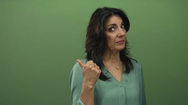 Latin kökenli orta yaşlı bir kadın yeşil bir stüdyoda parmak ucunda, kaş kaldırarak yan bakıyor; şüphe.