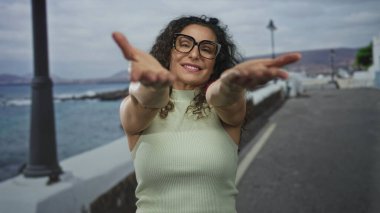 Kıvırcık saçlı, kolsuz üst ve siyah çerçeveli gözlüklü bir kadın gülümser ve deniz kenarında kollarını uzatır..