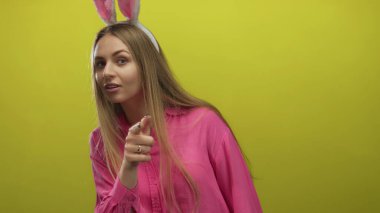 Tavşan kulaklı bir kadın, pembe gömlekli, sarı bir arka plana neşeli bir şekilde işaret ediyor. Genç, kaygısız bir havayla eğlenceli bir paskalya teması öneriyor..