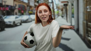 Şehir merkezinde futbol topunu tutan kadın canlı şehir ortamında, etkileyici jestler ve kendinden emin tavırları sergiliyor..