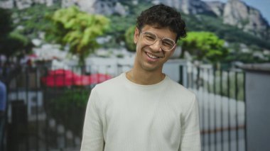 Gözlüklü ve görünür dişli bir adam gün ışığı altında taş binanın önünde gülümser. Mutluluk..