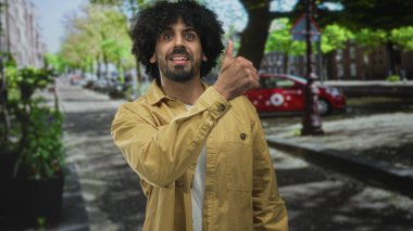 Sarı ceketli kıvırcık saçlı bir adam, gülümseyerek ve başparmak hareketi göstererek kentteki caddede baş parmağını kaldırıyor..