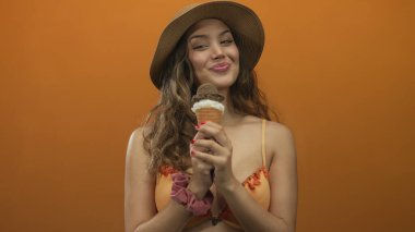 Bikinili bir kadın canlı turuncu arka planda, neşeli bir ifadeyle ve şık bir şapkayla dondurmadan hoşlanıyor..