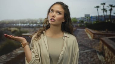 Parkta el hareketleriyle hareket eden kadın, bulutlu bir gökyüzünün altında bitki örtüsü ve palmiye ağaçlarının yer aldığı yeşil bulanık arka plana inanmadığını ifade ediyor..