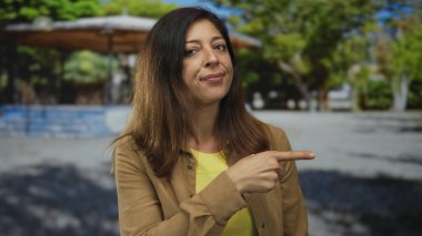 Parkta parkı işaret eden bir kadın, güneşli yeşil ağaçların ve gölgeli yolların ortasında ince bir gülümseme ile omzunun üzerinden bakarken;.