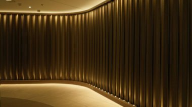 Dikey kıvrımları olan kıvrımlı perde duvarı ve sıcak altın ışıklandırması, hafifçe odağını kaybetmiş iç koridor; arka plan arka plan telif alanı sakinliği.