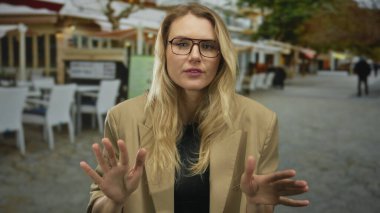 Şehir merkezindeki bir restoranın terasında sakince el kol hareketi yapan gözlüklü ve sarı saçlı bej ceketli bir kadın..
