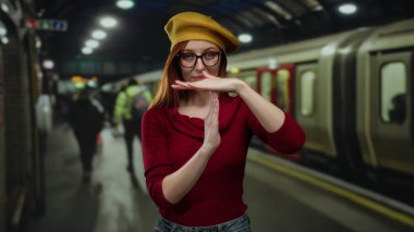 Tren istasyonunda sarı bereli, kırmızı kazaklı ve arka planda insanlar olan bir kadın..