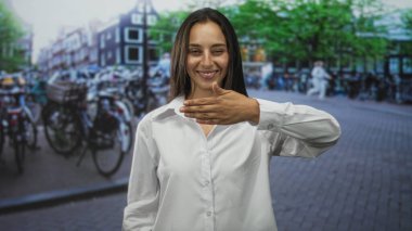 Ağzı eliyle kaplayan ve kaldırım taşı bir sokakta bisiklet ve binalarla gülümseyen bir kadın..