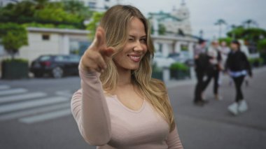 Sarışın kadın göz kırpıyor ve kameraya işaret ediyor yoğun sokak yaya geçidinde gün ışığında; şakacı flörtleşme.