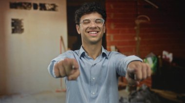 Gözlüklü genç adam bir inşaat alanındaki kamerayı işaret ediyor; dostça bir davet..