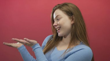 Açık mavi kazaklı kadın stüdyoda avuçlarını kırmızı arkaplan karşısında kameraya gösteriyor..