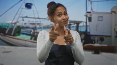 Kadın, iskele tarafındaki balıkçı teknelerinin yanındaki kamerayı işaret ediyor. Önlük ve uzun kollu bluz giyiyor..