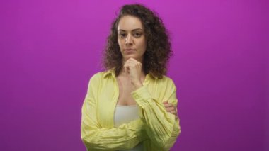 Stüdyoda eflatun arkaplan karşısında sarı gömlekli, çeneye dokunan genç İspanyol kadın..