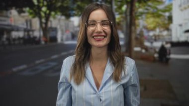 Gözlüklü ve açık mavi tişörtlü genç İspanyol kadın gülümsüyor ve güneşli bir caddeyi işaret ediyor; coşku.