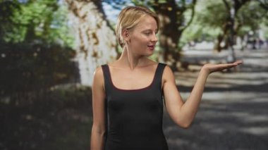 Siyah kolsuz tişörtlü sarışın kadın avucunu kaldırıyor ve ağaçlarla çevrili caddeye bakıyor; oyunculuk daveti.