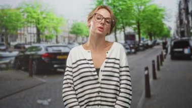 Sarışın kadın arabanın anahtarlarını tutuyor ve sokakta durup el kaldırıyor, çizgili hırka ve gözlük giyiyor, anahtarlığa diğer eliyle basıyor; tereddüt mahremiyeti.