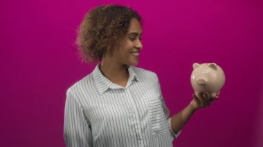 Domuz kumbarası tutan ve gülümseyen bir kadın, eflatun stüdyoda çizgili gömlek giyiyor; tasarruf bütçesi.