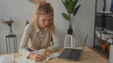 Masa başı çalışma sahnesinde dizüstü bilgisayarın yanında kalem tutan kadın sarışın yoğunluğunu gösteriyor..