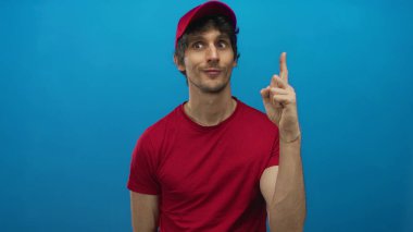 Kırmızı şapkalı ve gömlekli genç teslimatçı, mavi stüdyoda parmağını yukarı kaldırıyor. Düşünceli bir ifadeyle..