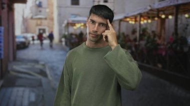 Genç ispanyol adam sokakta açık restoran terası ile dururken kamerayı işaret ediyor; davet bağlantısı.