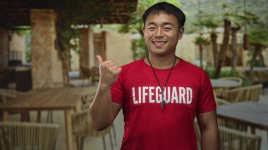 Kırmızı cankurtaran tişörtlü adam terasta duruyor. Islık kollu ve kolları açık gülümsüyor. Huzur..