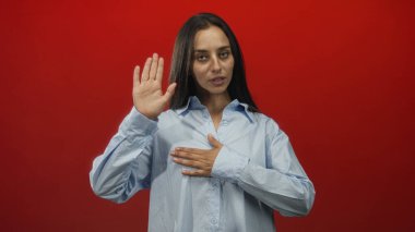 Kadın elini kırmızı stüdyoda kameraya kaldırırken diğer elini yemin işareti olarak göğsüne koyuyor..