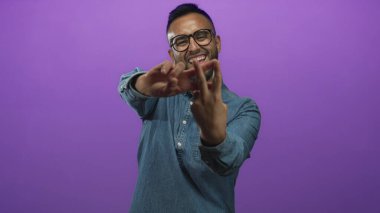 Gözlüklü ve kot gömlekli genç adam barış işareti yapıyor ve mor bir stüdyoda gülümsüyor; neşeli bir kutlama..