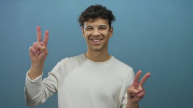 Mavi stüdyoda gülümseyen ve barış işareti yapan gözlüklü genç İspanyol adam iyimserlik.