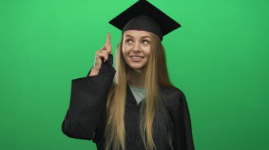 Mezuniyet üniformalı genç beyaz kadın yeşil arka planda kutlama yapıyor, neşeli bir ifadeyle, başarıyı ve başarıyı sembolize ediyor..