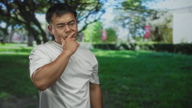 Yeşil ağaçlar ve çimlerle çevrili parktaki adam çenesine dokunuyor. Düşünceli bir hareket..