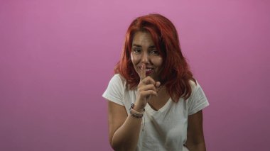 Sessiz kalmak için parmakları dudakları olan bir kadın pembe bir stüdyoda kameraya karşı sessiz kalıyor beyaz t-shirt ve bilezikler takıyor; şakacı gizlilik.