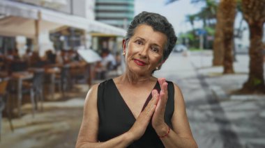 Şehir merkezinde açık kafeleri ve palmiye ağaçları olan canlı bir caddede el çırpan gri saçlı yaşlı bir kadın..