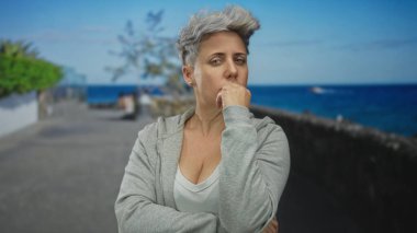 Gri kapüşonlu kısa gri saçlı kadın, sokakta çene jesti el ele durur; sessiz tefekkür.