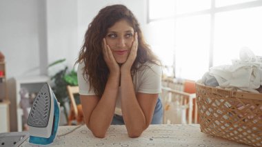 Elleri yanaklı kadın ütü masasına yaslanmış evde demir ve çamaşır sepetiyle; ev konforu.