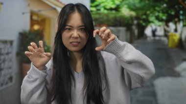 Yeşilliklerle dolu bir sokakta neşeli bir jest yapan, günlük kıyafetler giyen ve enerjik bir şekilde gülümseyen, hayat dolu bir şehir ortamında güven ve neşe saçan bir kadın..