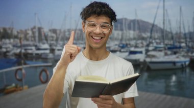 Gözlüklü genç İspanyol adam açık bir kitap tutuyor ve tahta bir iskelede okurken parmakla işaret ediyor; merak.